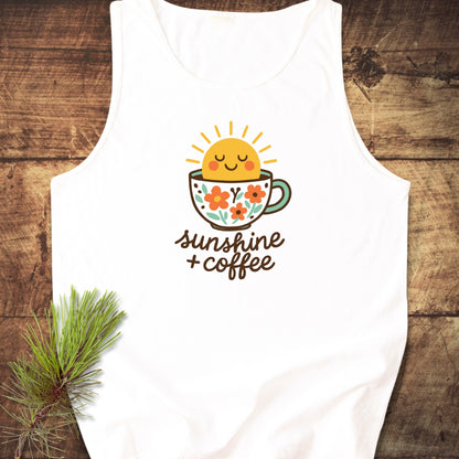 Sunshine And Coffee Tank Top featuring a smiling sun in a floral coffee cup graphic with sunshine + coffee text, displayed flat on wood with a pine branch accent.