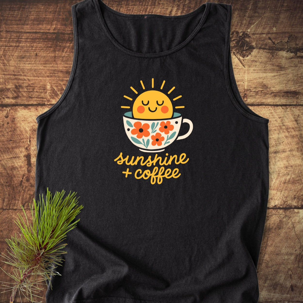 Sunshine And Coffee Tank Top displayed on a wooden surface, featuring a cheerful sun in a floral coffee cup graphic. Includes sunshine + coffee text. Accented by a pine branch beside it.