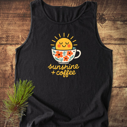 Sunshine And Coffee Tank Top displayed on a wooden surface, featuring a cheerful sun in a floral coffee cup graphic. Includes sunshine + coffee text. Accented by a pine branch beside it.