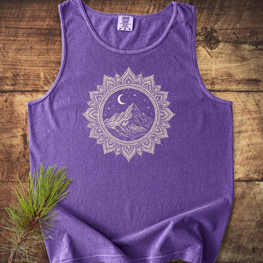Mountain Mandala Tank Top: Purple, sleeveless Comfort Colors tank top with a mandala-style graphic of mountains under a crescent moon and stars, laid flat on wood with a pine branch accent.