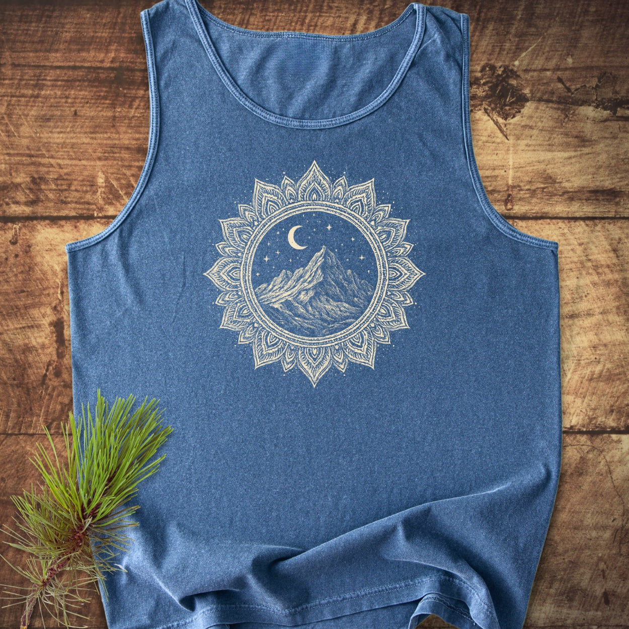 Mountain Mandala Tank Top features a heathered design with a circular mandala graphic of a mountain peak under a crescent moon and stars, laid flat with a pine twig beside it.