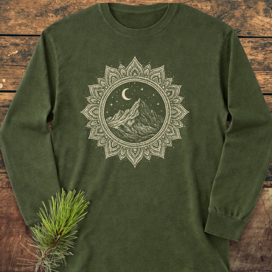 Mountain Mandala Long-Sleeve Tee featuring a hand-drawn mountain and moon graphic on olive-green fabric, laid flat on wood. Made from soft, 100% ring-spun cotton, offering a relaxed, comfortable fit.