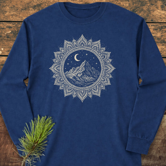 Mountain Mandala Long-Sleeve Tee featuring an intricate off-white mandala and mountain design on deep-blue cotton, laid on wooden surface with pine sprig accent, offering softness and a relaxed fit.