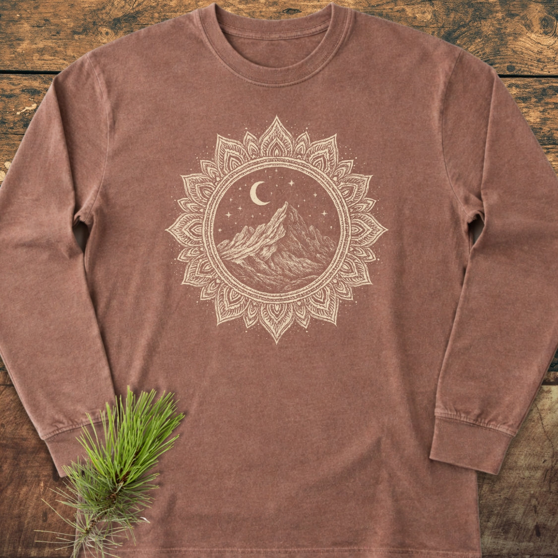 Mountain Mandala Long-Sleeve Tee displayed on wood, featuring a mandala graphic with mountains, crescent moon, and stars. Styled with a pine branch, highlighting its soft, garment-dyed 100% ring-spun cotton material.