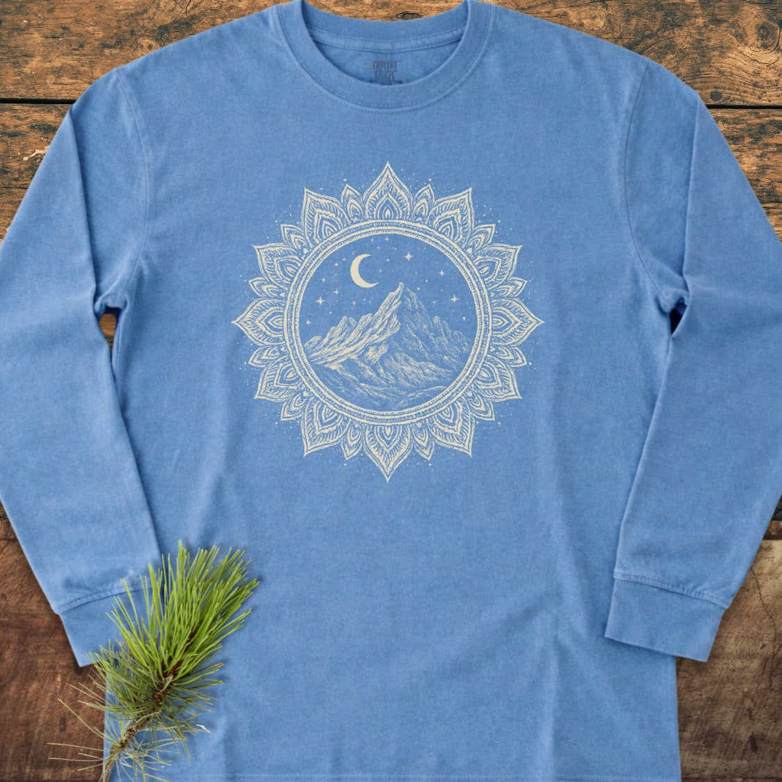 Mountain Mandala Long-Sleeve Tee laid flat on wood, showcasing a white mandala mountain graphic under moon and stars. Pine twig beside. Made from 100% ring-spun cotton for comfort and style.