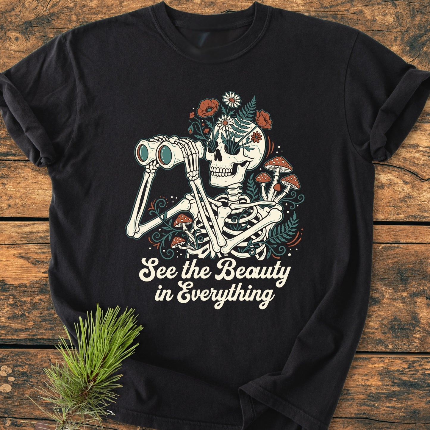 See The Beauty - Vintage Wash Tee featuring a skeleton with binoculars, flowers, and mushrooms, printed on a relaxed-fit, soft-washed garment-dyed shirt, laid on rustic wood with pine sprig accent.