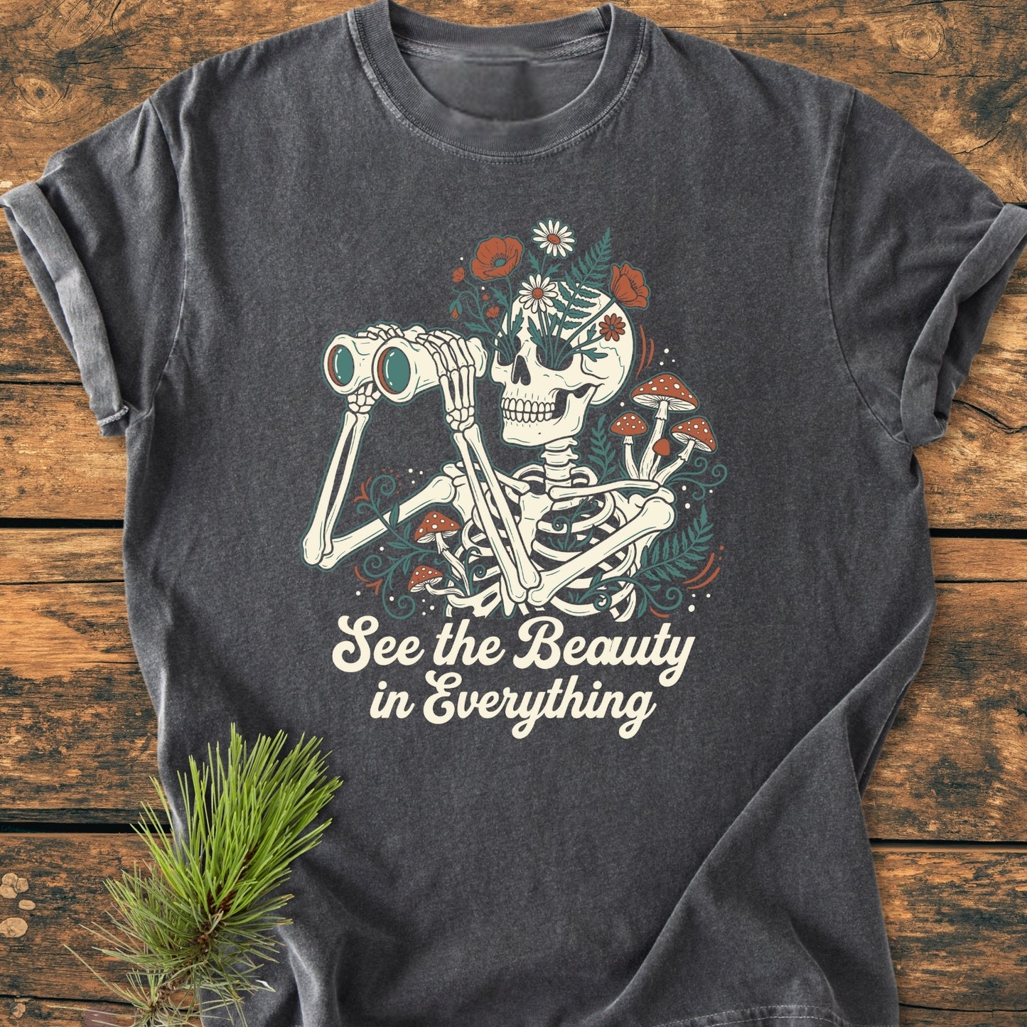 See The Beauty - Vintage Wash Tee features a whimsical skeleton with binoculars, wildflowers, and mushrooms, below the slogan “See the Beauty in Everything,” on a dark-gray, soft, relaxed-fit cotton tee.