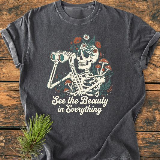 See The Beauty - Vintage Wash Tee features a whimsical skeleton with binoculars, wildflowers, and mushrooms, below the slogan “See the Beauty in Everything,” on a dark-gray, soft, relaxed-fit cotton tee.