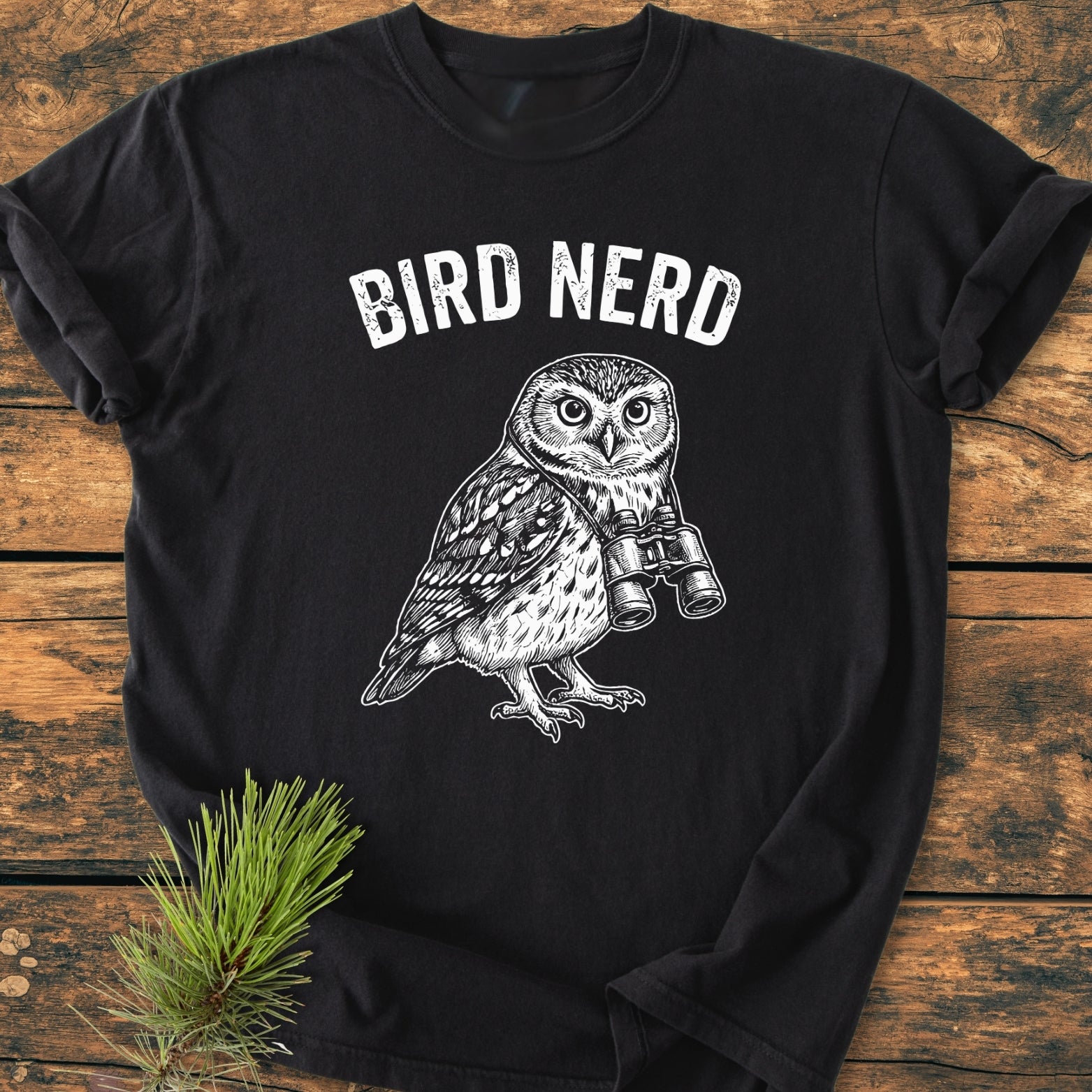 Bird Nerd Owl - Vintage Wash Tee featuring “BIRD NERD” text and owl with binoculars. Laid on wood, pine sprig beside. Soft, durable, relaxed fit, made of 100% ring-spun cotton.