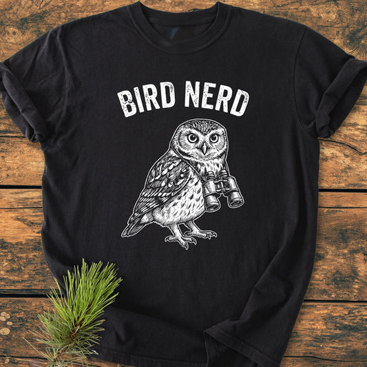 Bird Nerd Owl - Vintage Wash Tee featuring “BIRD NERD” text and owl with binoculars. Laid on wood, pine sprig beside. Soft, durable, relaxed fit, made of 100% ring-spun cotton.