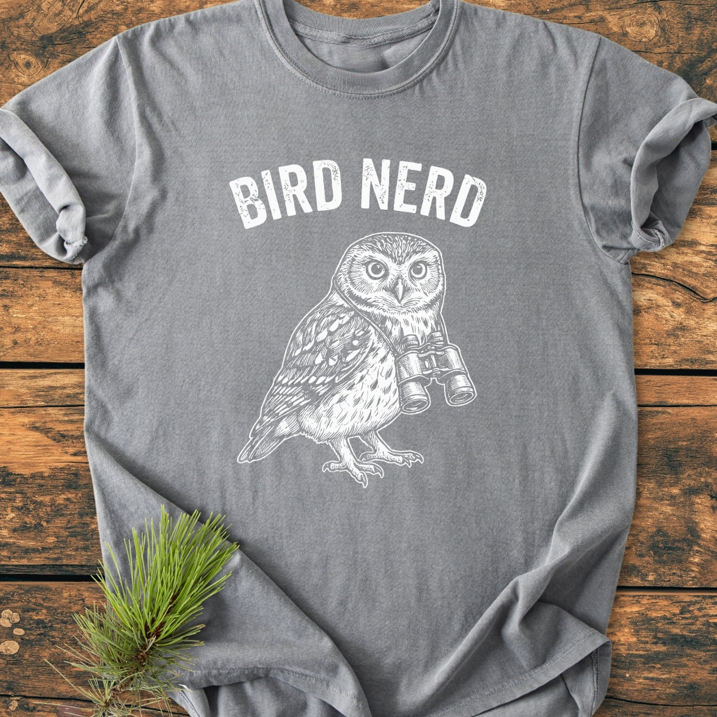 Heather-gray Bird Nerd Owl - Vintage Wash Tee features a white BIRD NERD print and owl graphic with binoculars, laid on wooden surface with pine sprig.