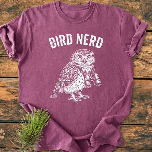 Bird Nerd Owl - Vintage Wash Tee features an owl with binoculars and BIRD NERD text. Laid on wood, it has rolled sleeves and a pine sprig accentuating its rustic charm.
