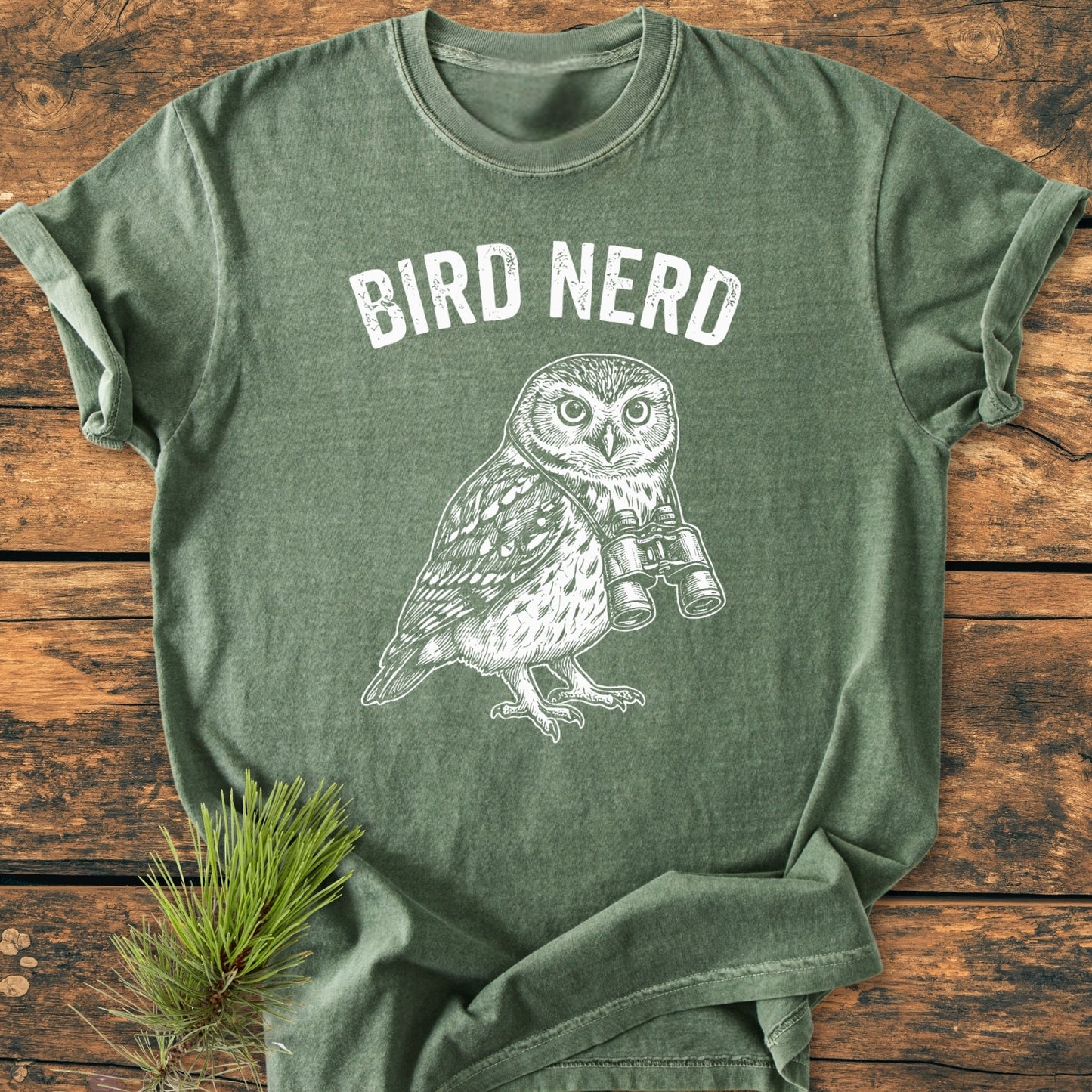 The Bird Nerd Owl - Vintage Wash Tee features a BIRD NERD print above an owl with binoculars, displayed flat on a wooden surface, highlighting its relaxed fit and soft-washed, ring-spun cotton fabric.
