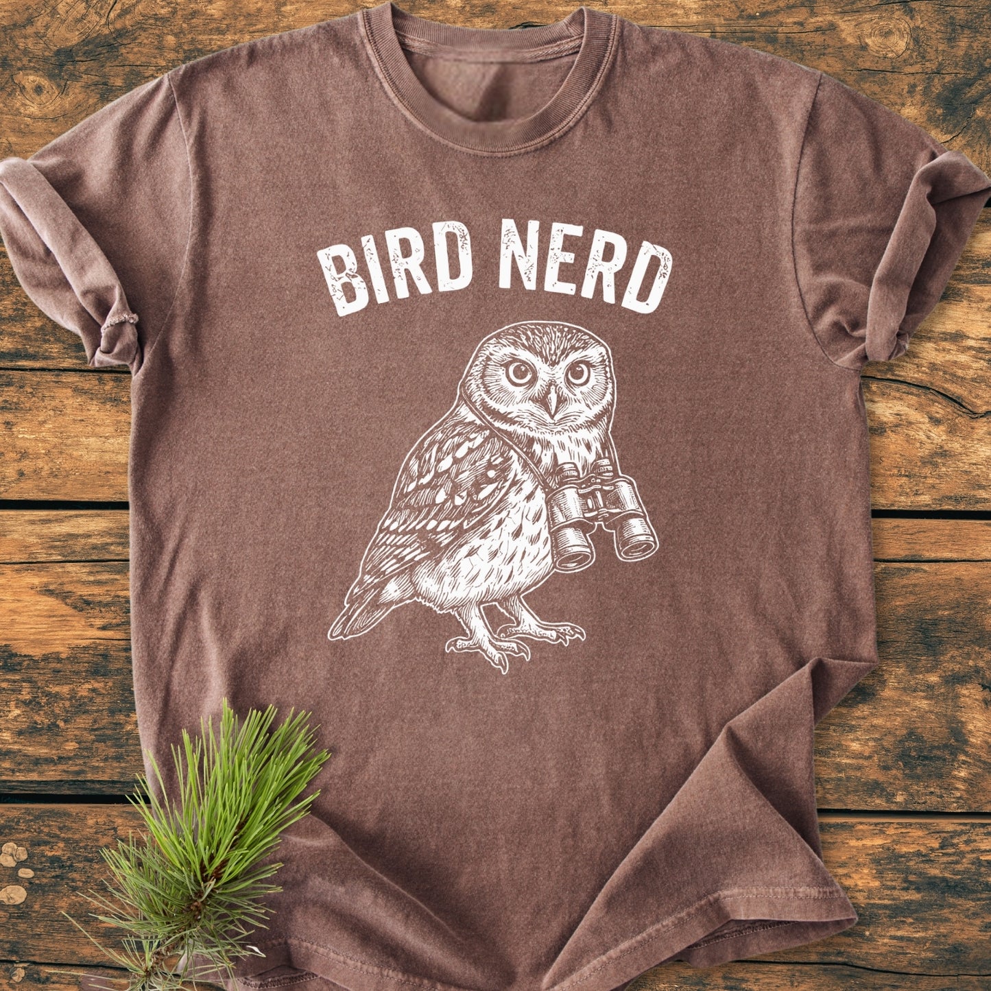 Bird Nerd Owl - Vintage Wash Tee featuring BIRD NERD and an owl with binoculars, laid flat on wood. Styled with a pine bough, showcasing its relaxed fit and durable stitching.