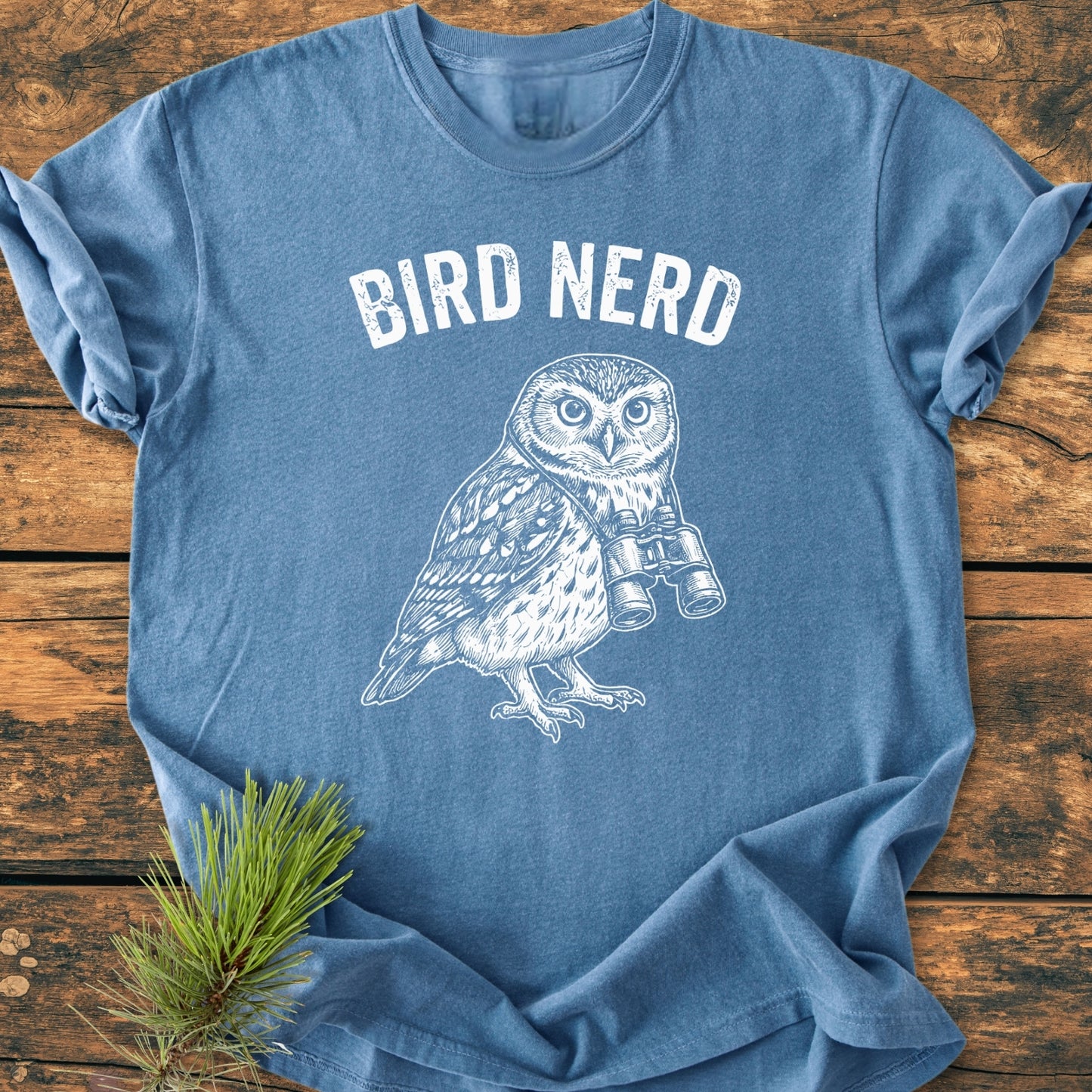 Bird Nerd Owl - Vintage Wash Tee features a white BIRD NERD print and an owl with binoculars on a blue T-shirt, laid on wood with a pine sprig.