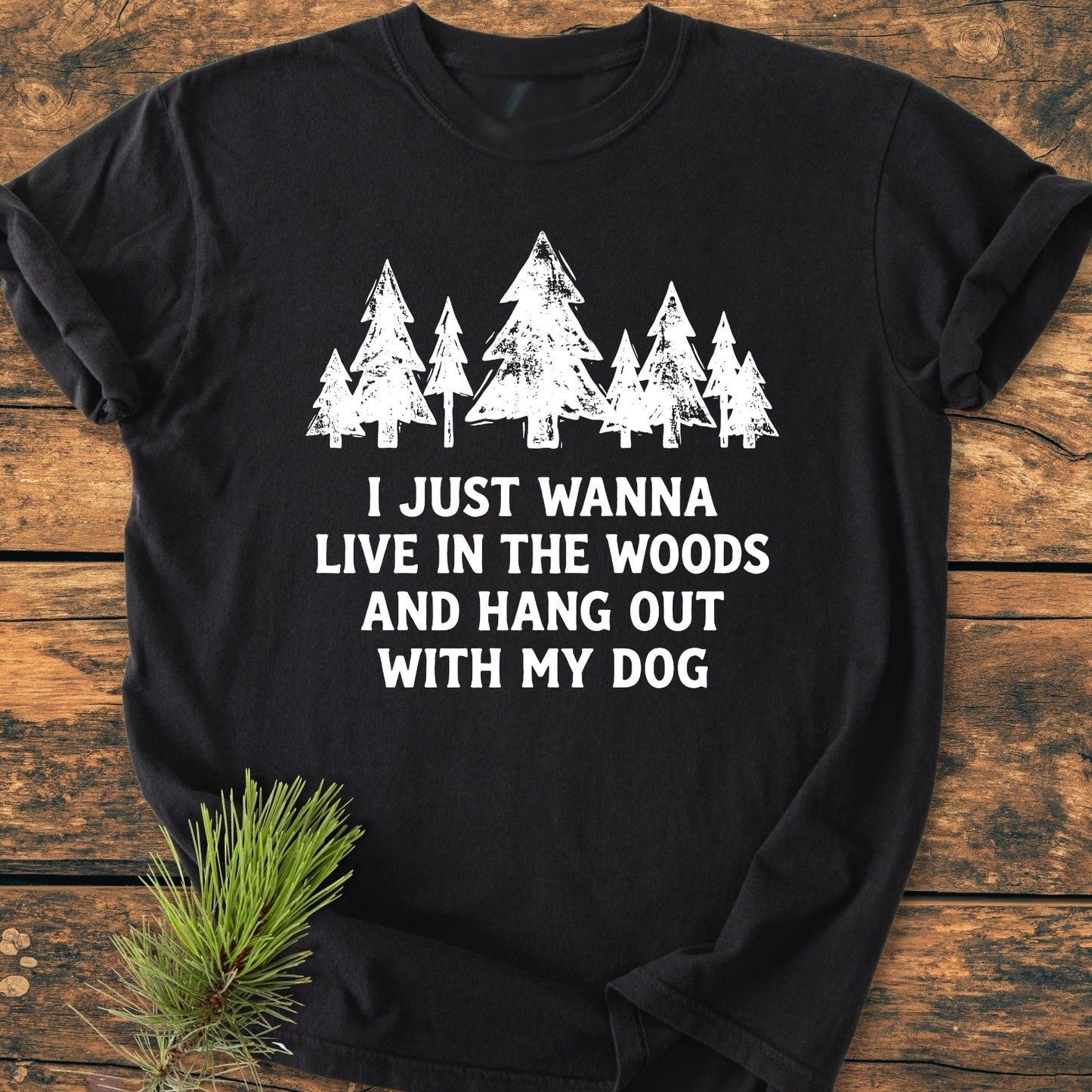 Live In The Woods - Vintage Wash Tee: Black T-shirt with white pine trees and text; displayed flat with a pine branch beside it, highlighting its relaxed fit and soft, garment-dyed fabric.