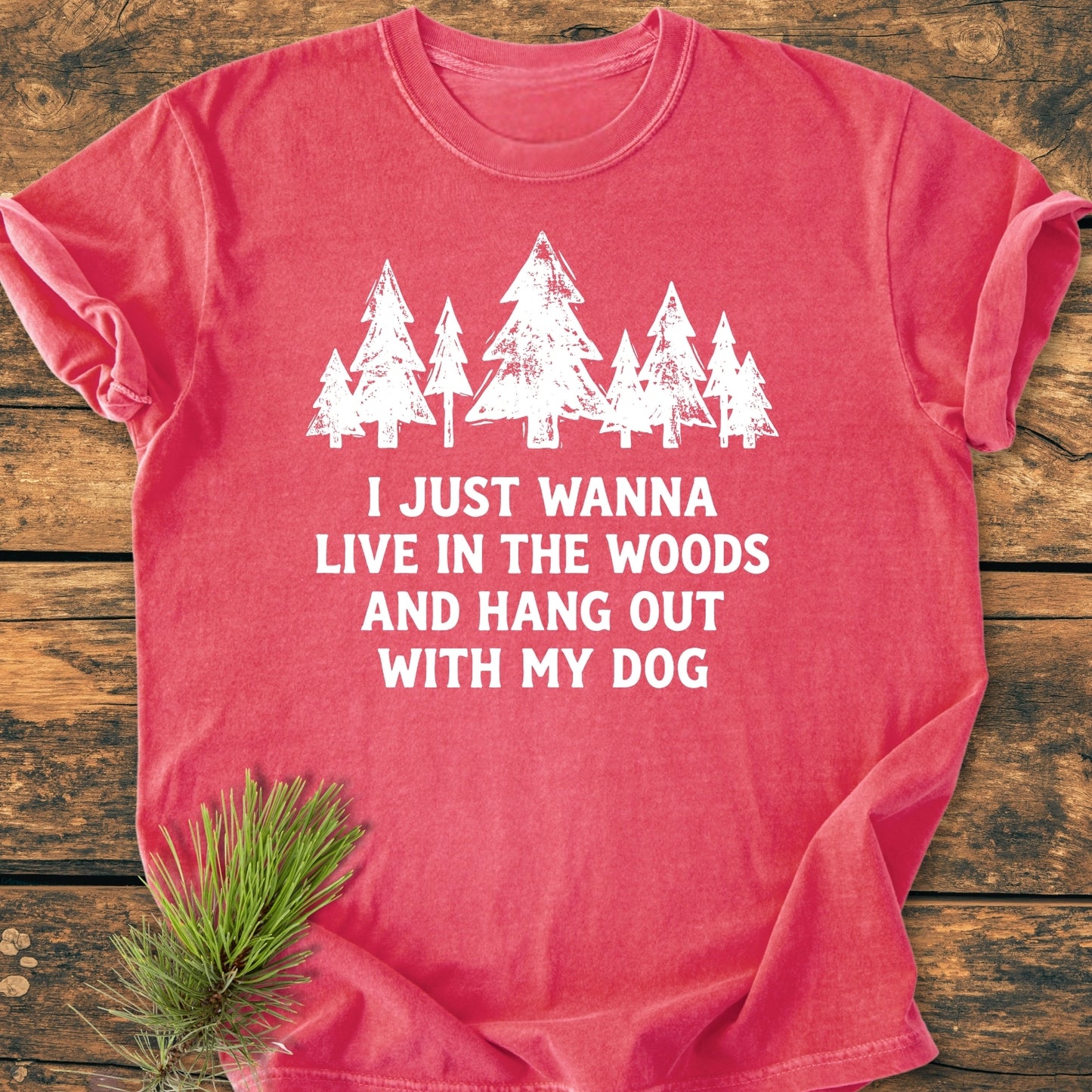 Live In The Woods - Vintage Wash Tee features a white pine tree graphic and slogan on a red-coral shirt, laid flat on wood, with a pine sprig beside it.