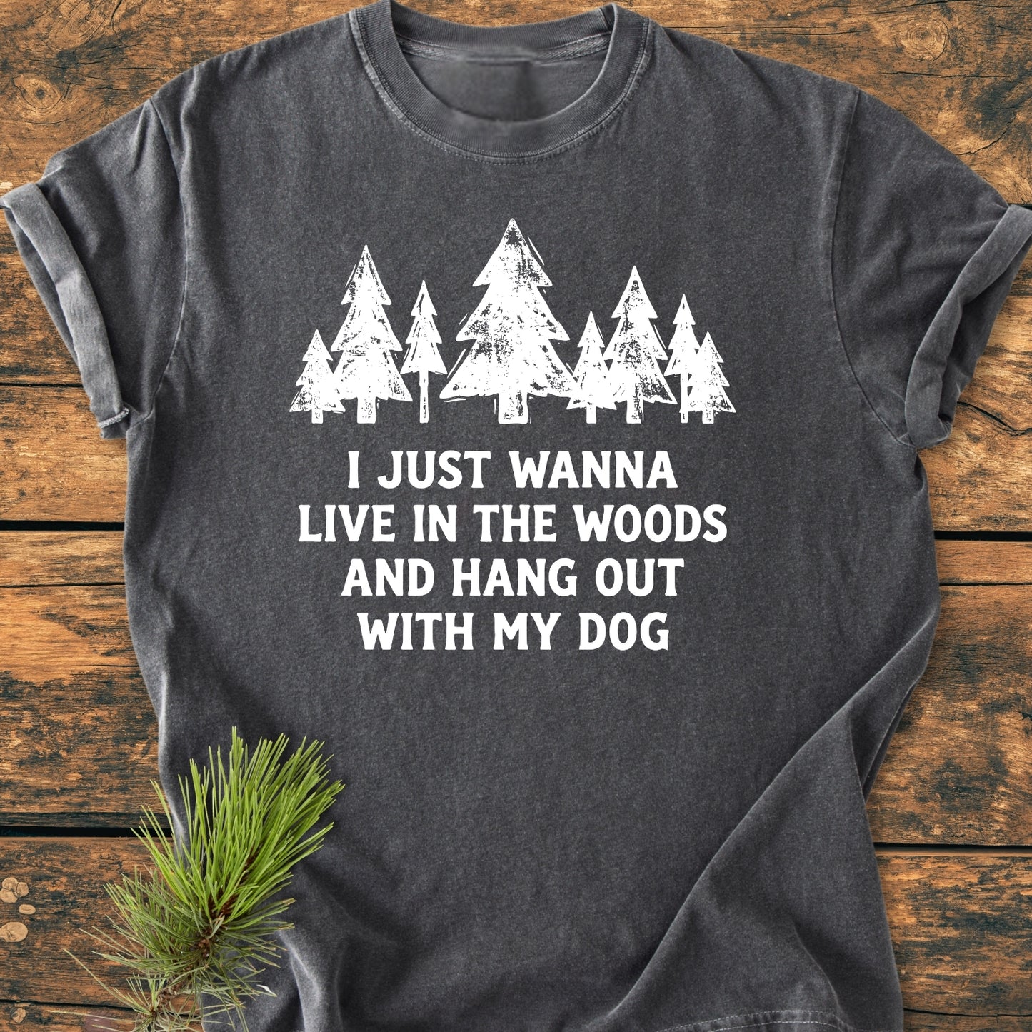 Live In The Woods - Vintage Wash Tee features a row of white pine trees and text on a charcoal-gray T-shirt, laid flat with a pine sprig beside it on a wooden surface.