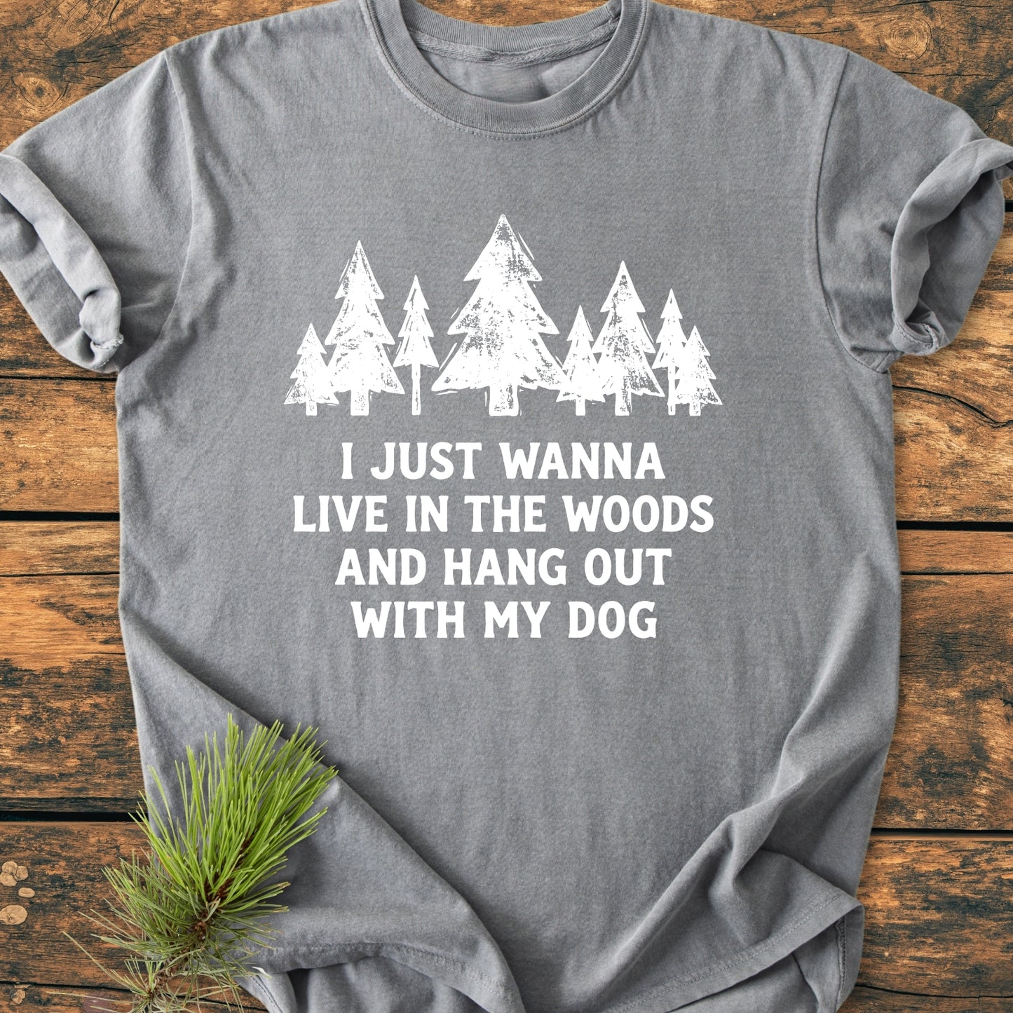 Live In The Woods - Vintage Wash Tee featuring a row of white stylized pine trees and a nature-inspired quote, displayed on a rustic wooden surface with a pine sprig accent.