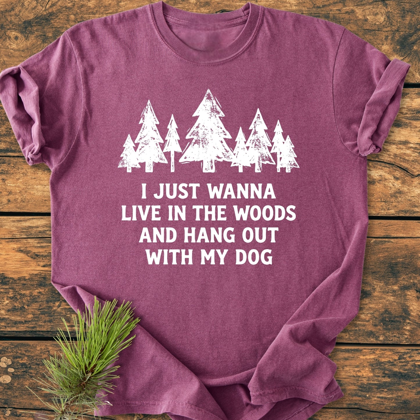 Live In The Woods - Vintage Wash Tee featuring white stylized pine trees and slogan, laid out on a wooden surface with a decorative pine branch beside, showcasing its relaxed fit and soft-washed fabric.