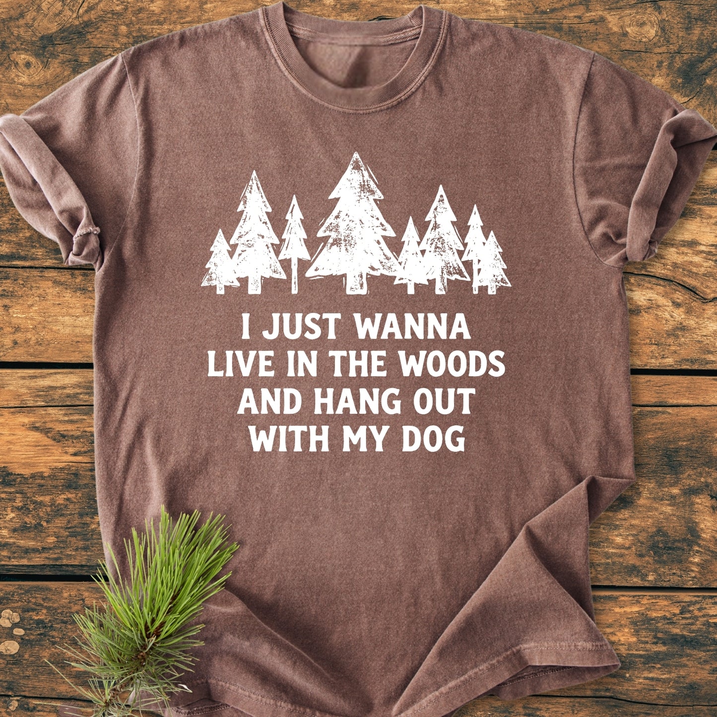 Live In The Woods - Vintage Wash Tee: Brown cotton T-shirt with white stylized pine trees and slogan, laid flat on wood surface, complemented by an evergreen sprig for styling.