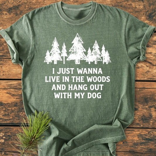 Live In The Woods - Vintage Wash Tee on wooden surface, features white screen-printed pine trees and text. Accompanied by a pine sprig, highlighting the shirt's forest-inspired design.