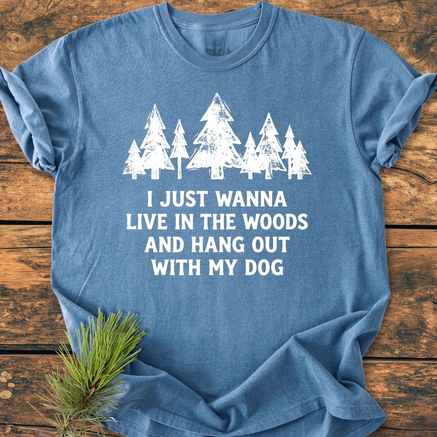 Live In The Woods - Vintage Wash Tee featuring white pine trees and text on soft, blue fabric, laid on wood. A pine sprig accents this relaxed, durable, cotton shirt, perfect for nature lovers.