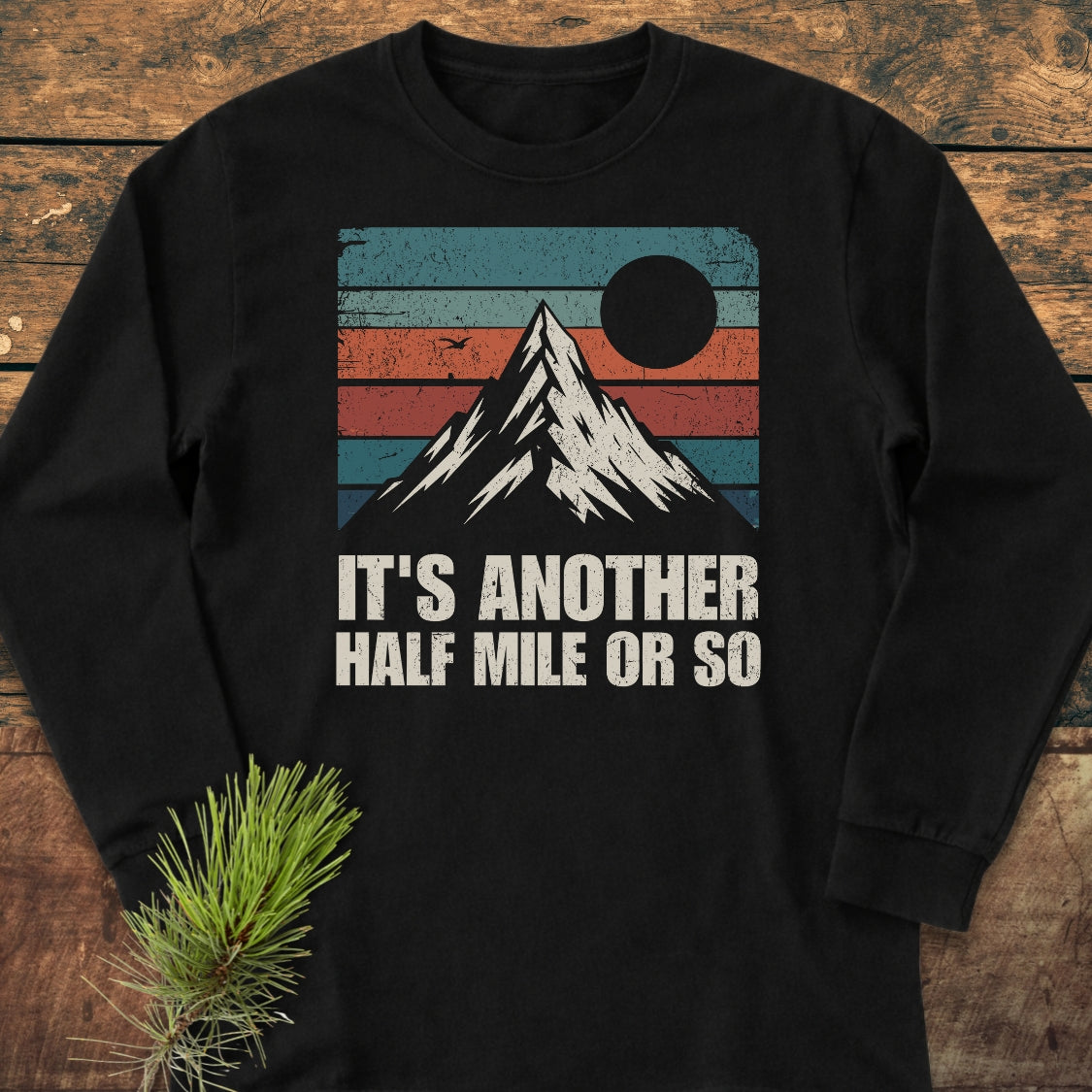It's Another Half Mile Or So Long-Sleeve Tee featuring a retro mountain graphic with teal and rust stripes, sun or moon, on black cotton, styled with a pine branch on a wooden surface.