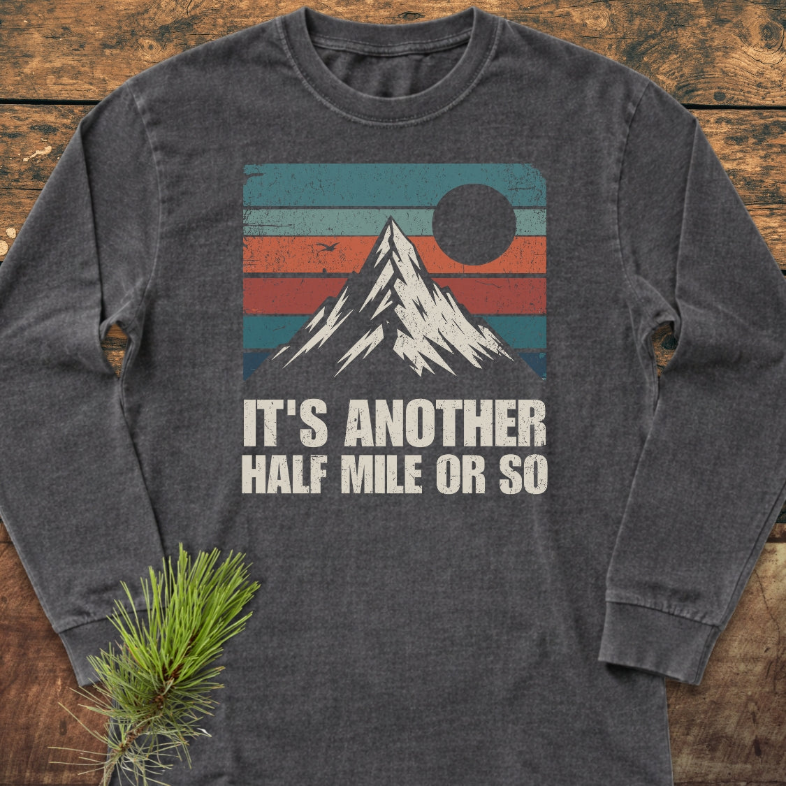 It's Another Half Mile Or So Long-Sleeve Tee featuring a retro mountain graphic with teal, orange, red, and blue stripes, laid on wood. Made from soft, 100% ring-spun cotton for a relaxed fit.
