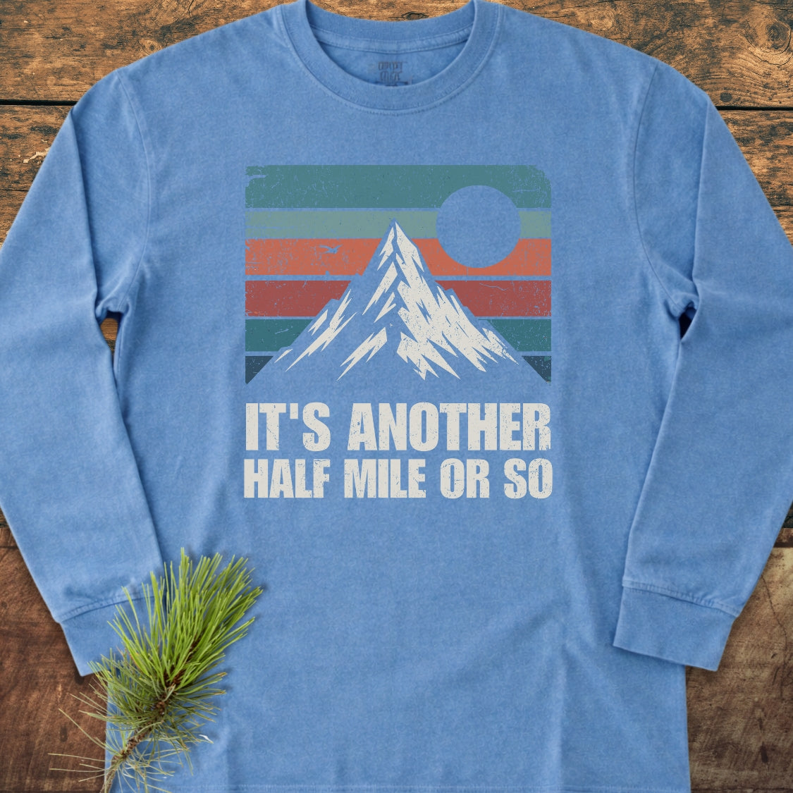 It's Another Half Mile Or So Long-Sleeve Tee features a retro graphic of a mountain with a sun, made from soft, garment-dyed 100% ring-spun cotton, laid on wooden surface.