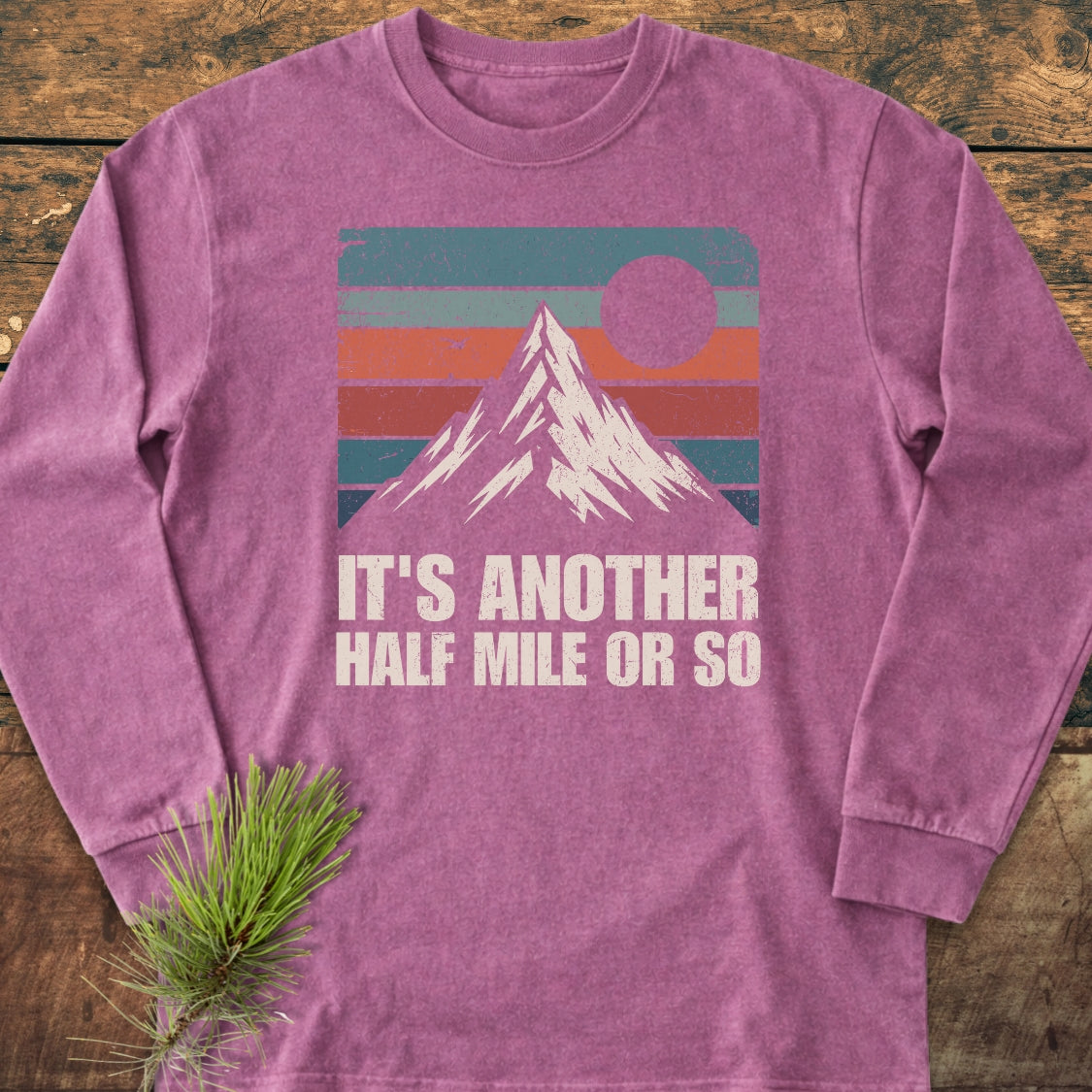 The It's Another Half Mile Or So Long-Sleeve Tee features a mountain graphic with sunset stripes, laid on wood. A pine sprig accents the soft, relaxed-fit, heathered-purple shirt.