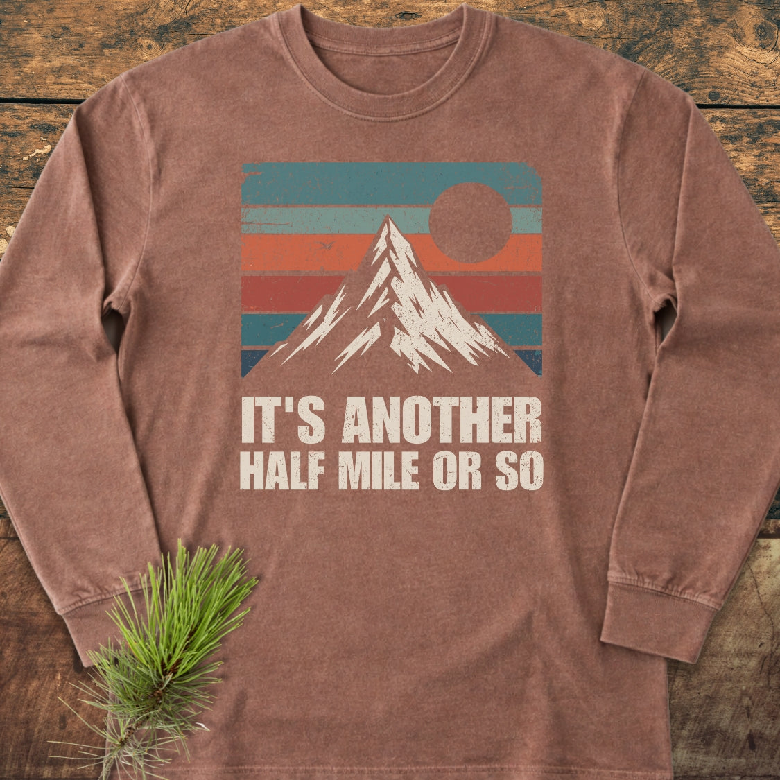 It's Another Half Mile Or So Long-Sleeve Tee features a retro mountain graphic with a slogan on soft, brown fabric, styled with a pine branch for a cozy outdoor aesthetic.