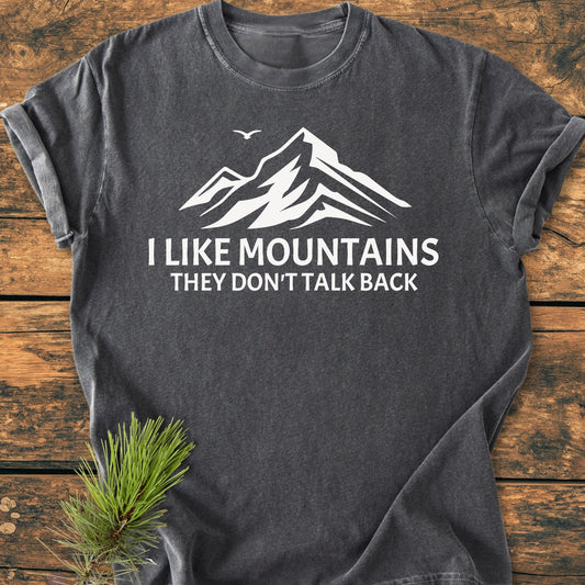 Charcoal-gray I Like Mountains - Vintage Wash Tee with white mountain illustration and slogan, laid flat on wood. Features include ring-spun cotton, soft-washed fabric, and relaxed fit. Pine sprig accent nearby.