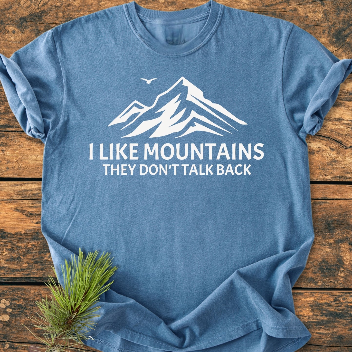 I Like Mountains - Vintage Wash Tee featuring a mountain silhouette and slogan on a heather-blue, short-sleeve design, displayed flat on wood with a pine sprig.