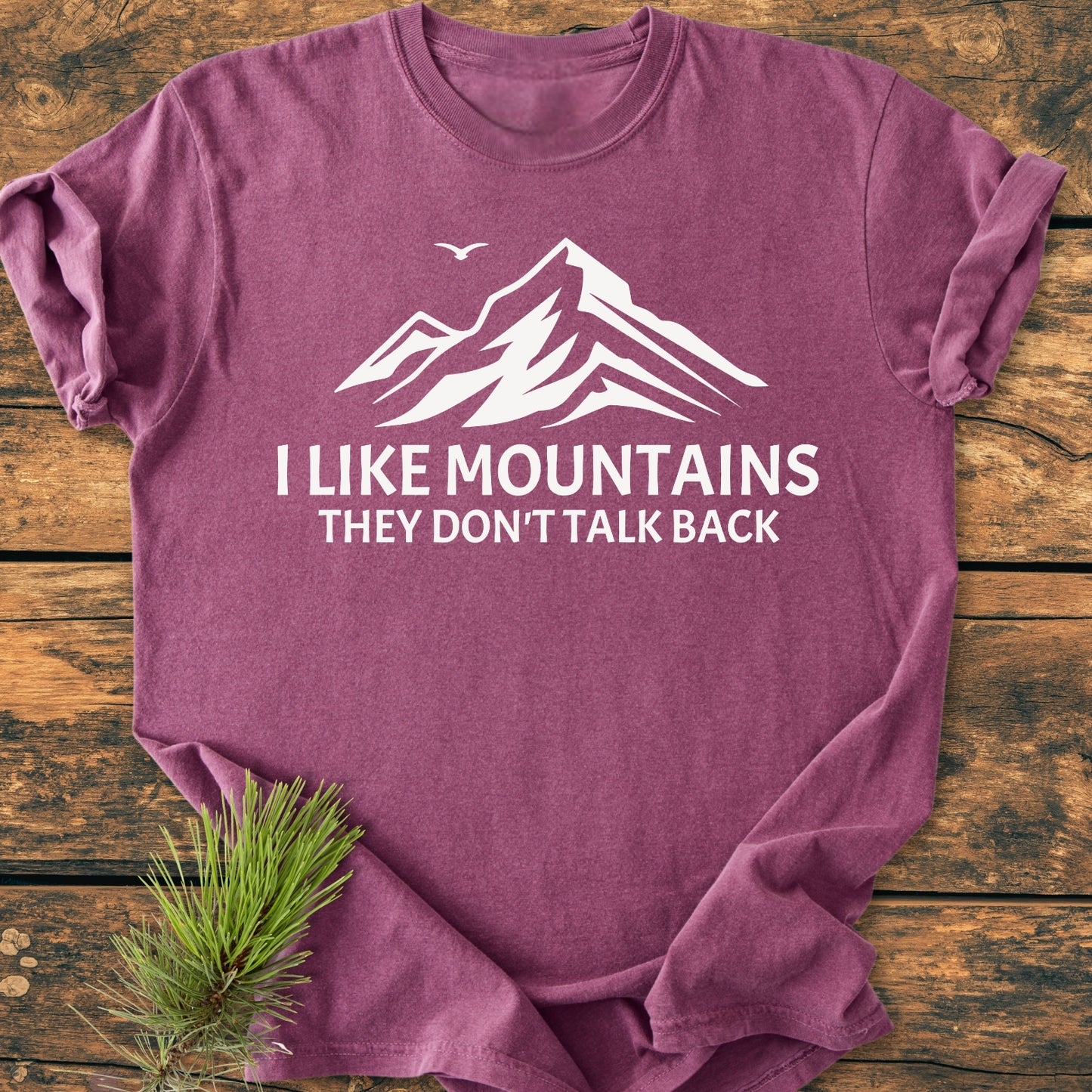 Heather-purple I Like Mountains - Vintage Wash Tee with a white mountain graphic and slogan, laid flat on wood with an evergreen sprig, showcasing its relaxed fit and durable stitching.