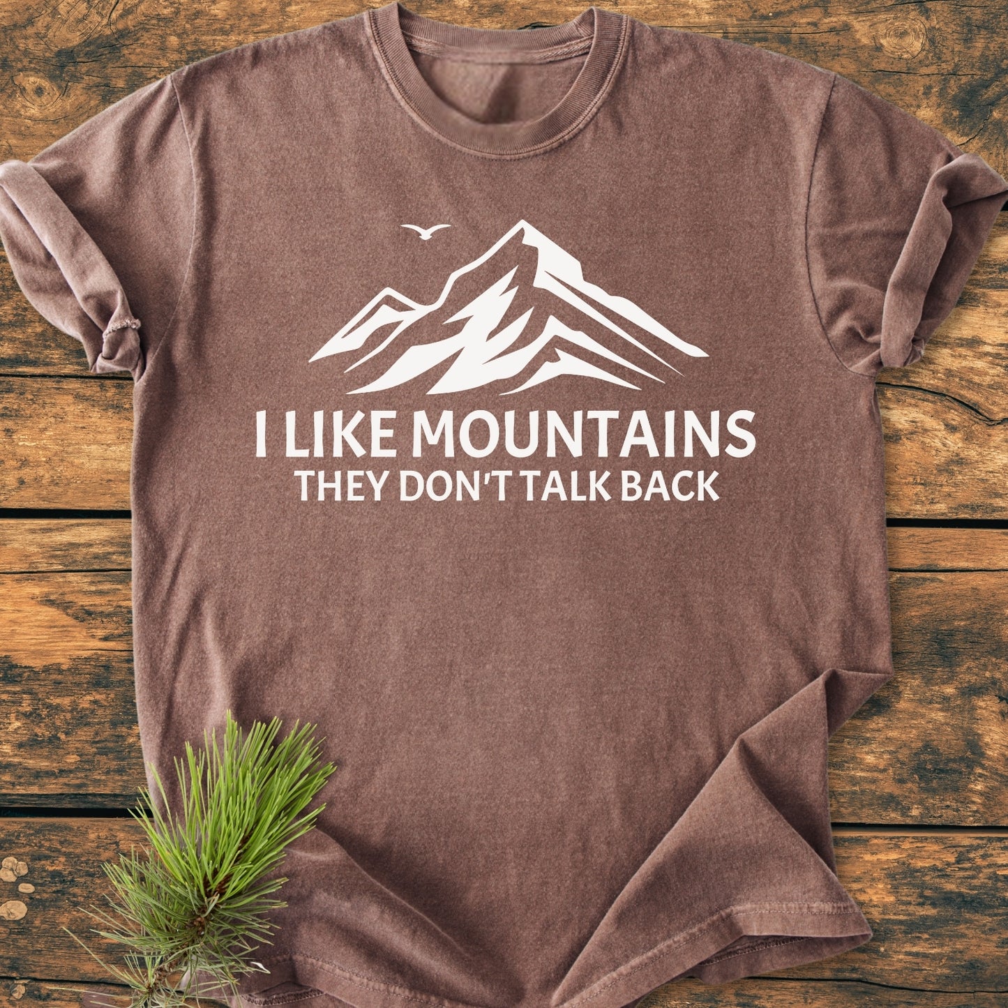 I Like Mountains - Vintage Wash Tee on rustic wood, showcasing a mountain graphic with bird silhouette and slogan. Features soft, garment-dyed fabric and relaxed fit for comfort.