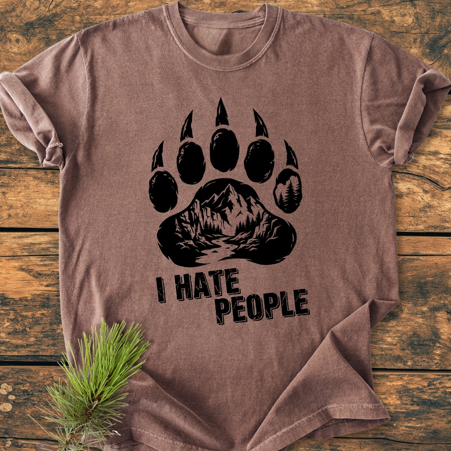I Hate People - Vintage Wash Tee featuring a bear paw print with a mountain-forest scene. Laid flat, showcasing its relaxed fit. Pine needles accentuate the garment's rustic charm.