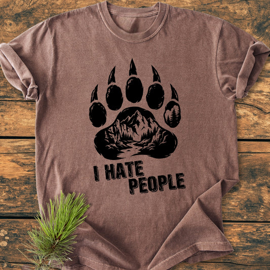 I Hate People - Vintage Wash Tee featuring a bear paw print with a mountain-forest scene. Laid flat, showcasing its relaxed fit. Pine needles accentuate the garment's rustic charm.