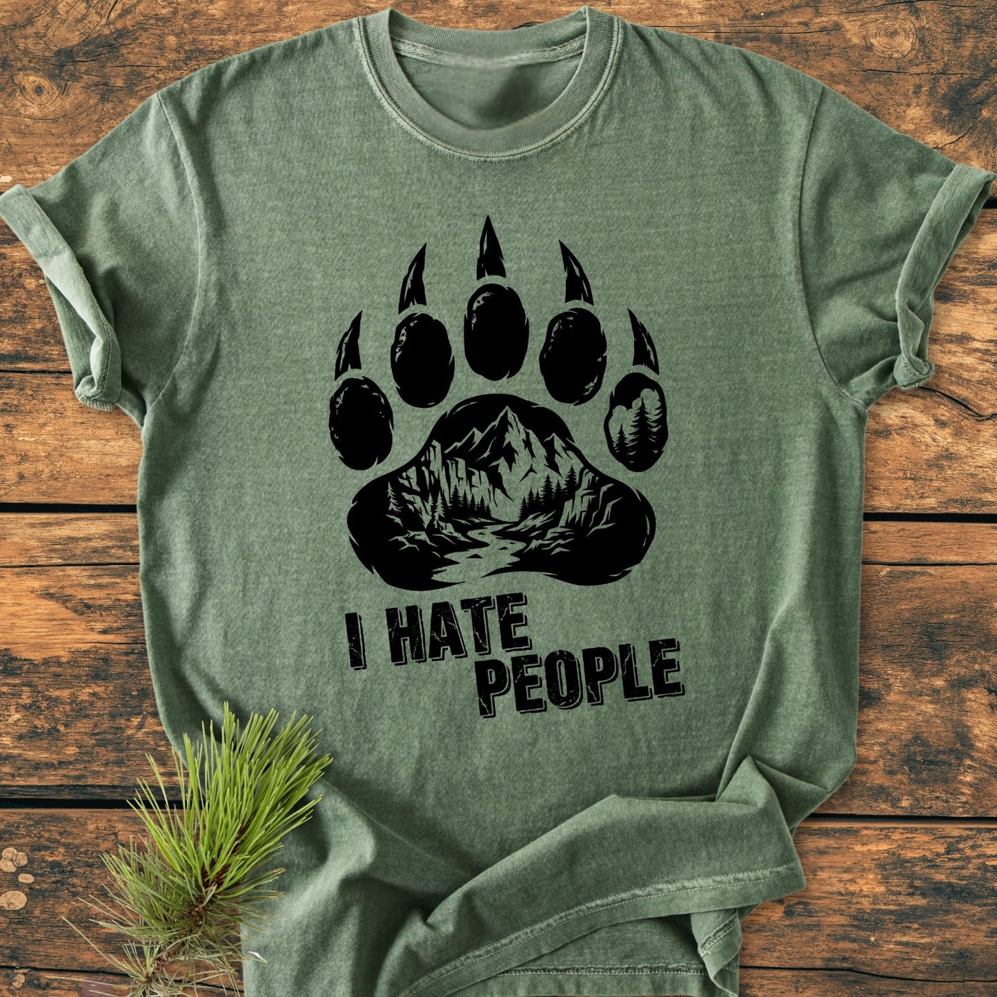 Heather-green I Hate People - Vintage Wash Tee displaying a bear-paw graphic with a mountain-and-forest scene, laid on a wooden surface. The tee features a relaxed fit and durable double-needle stitching.