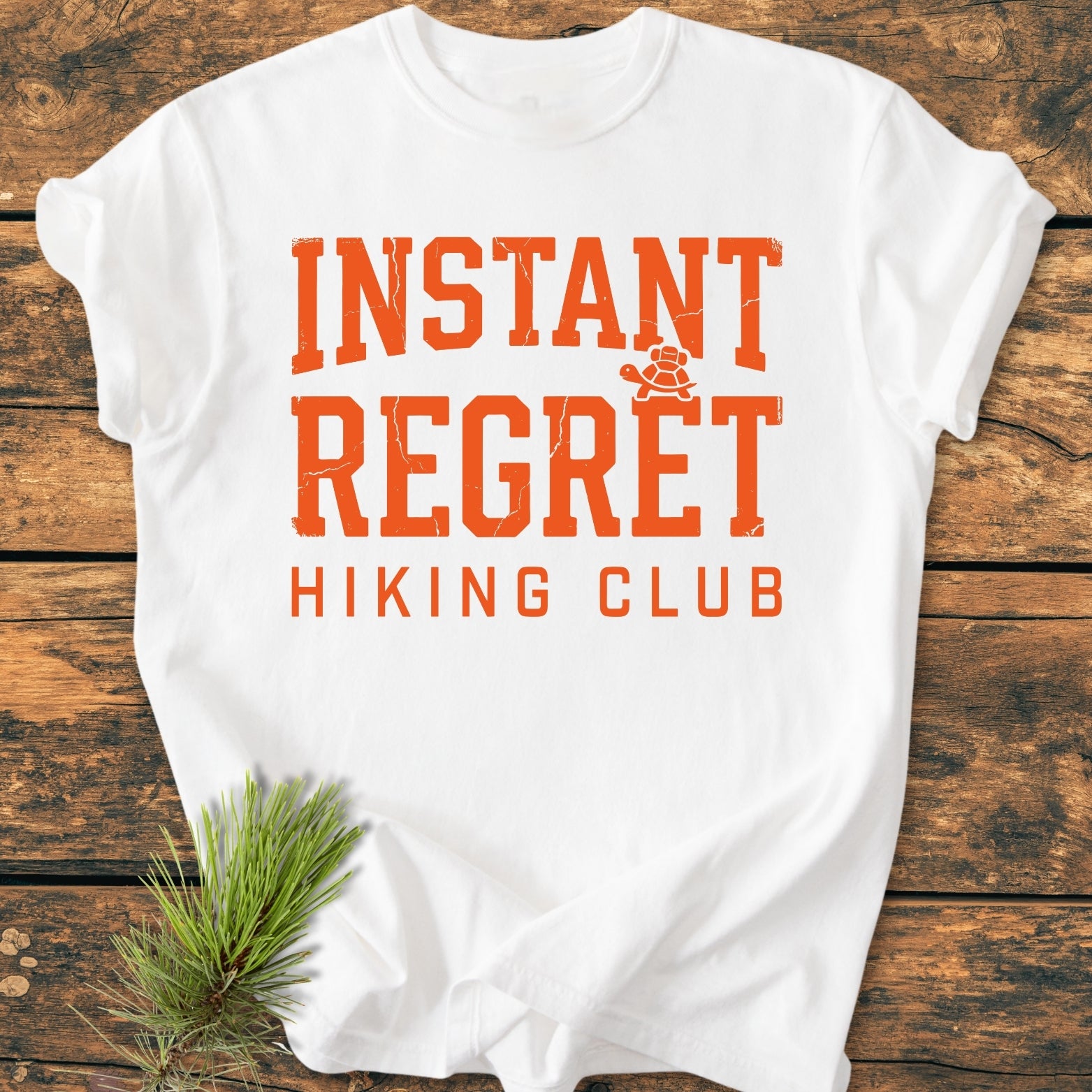 Instant Regret Hiking Club - Vintage Wash Tee displayed flat with bold distressed orange text and a turtle icon; set on rustic wood with a pine sprig, embodying an outdoorsy theme.