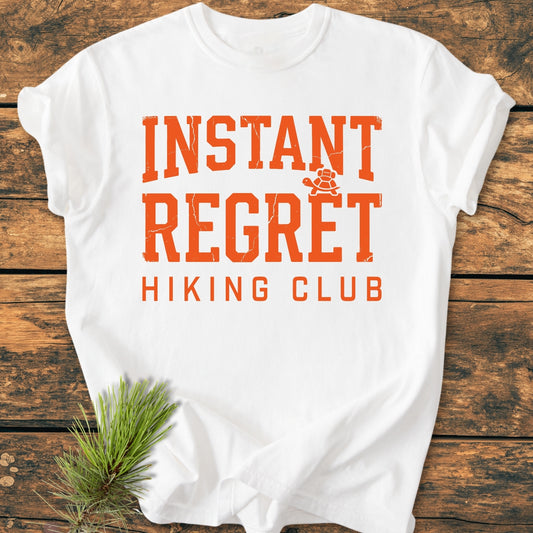 Instant Regret Hiking Club - Vintage Wash Tee displayed flat with bold distressed orange text and a turtle icon; set on rustic wood with a pine sprig, embodying an outdoorsy theme.