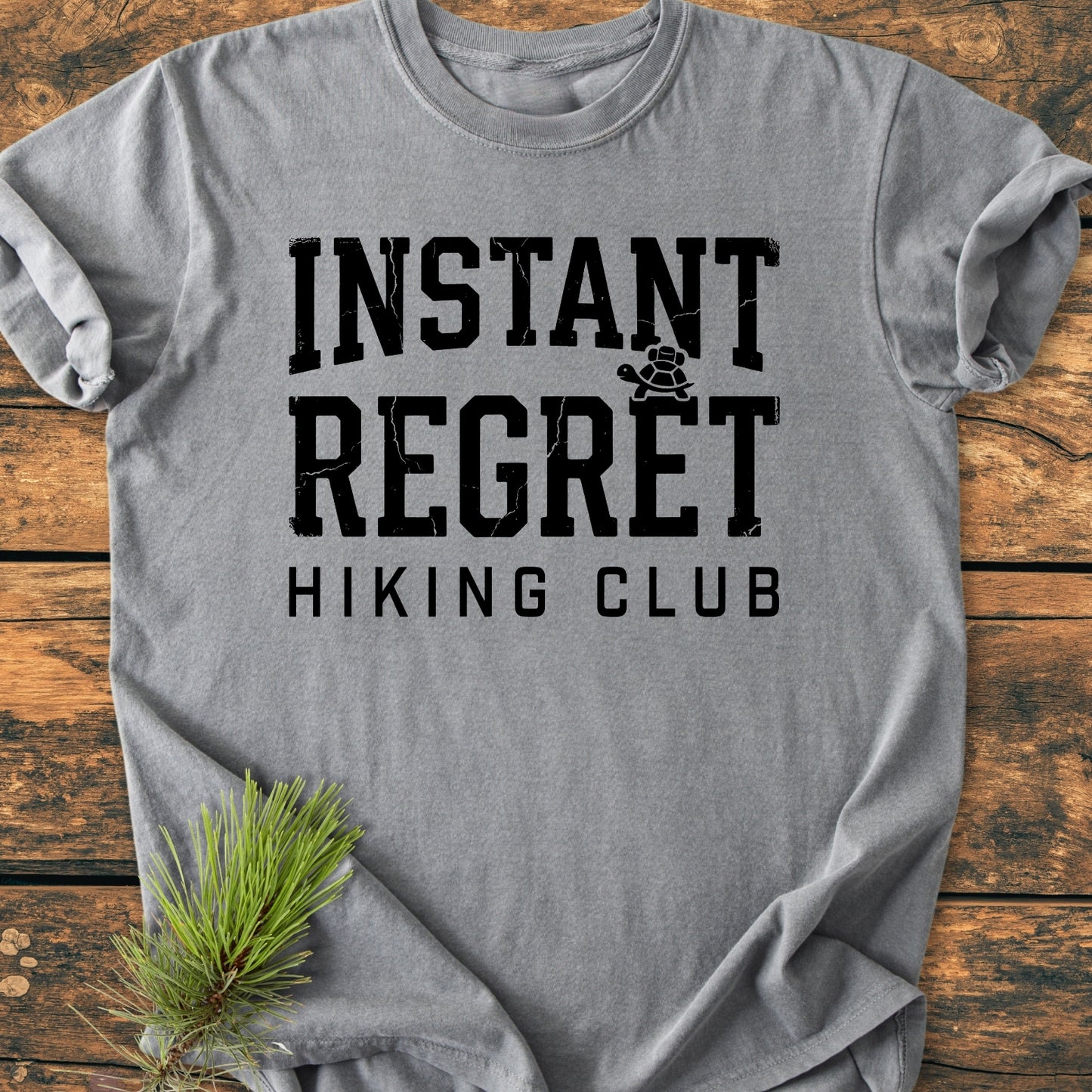 Instant Regret Hiking Club - Vintage Wash Tee features bold, distressed lettering with a turtle graphic on a soft, relaxed-fit light-gray T-shirt, styled with a pine sprig for a rustic touch.