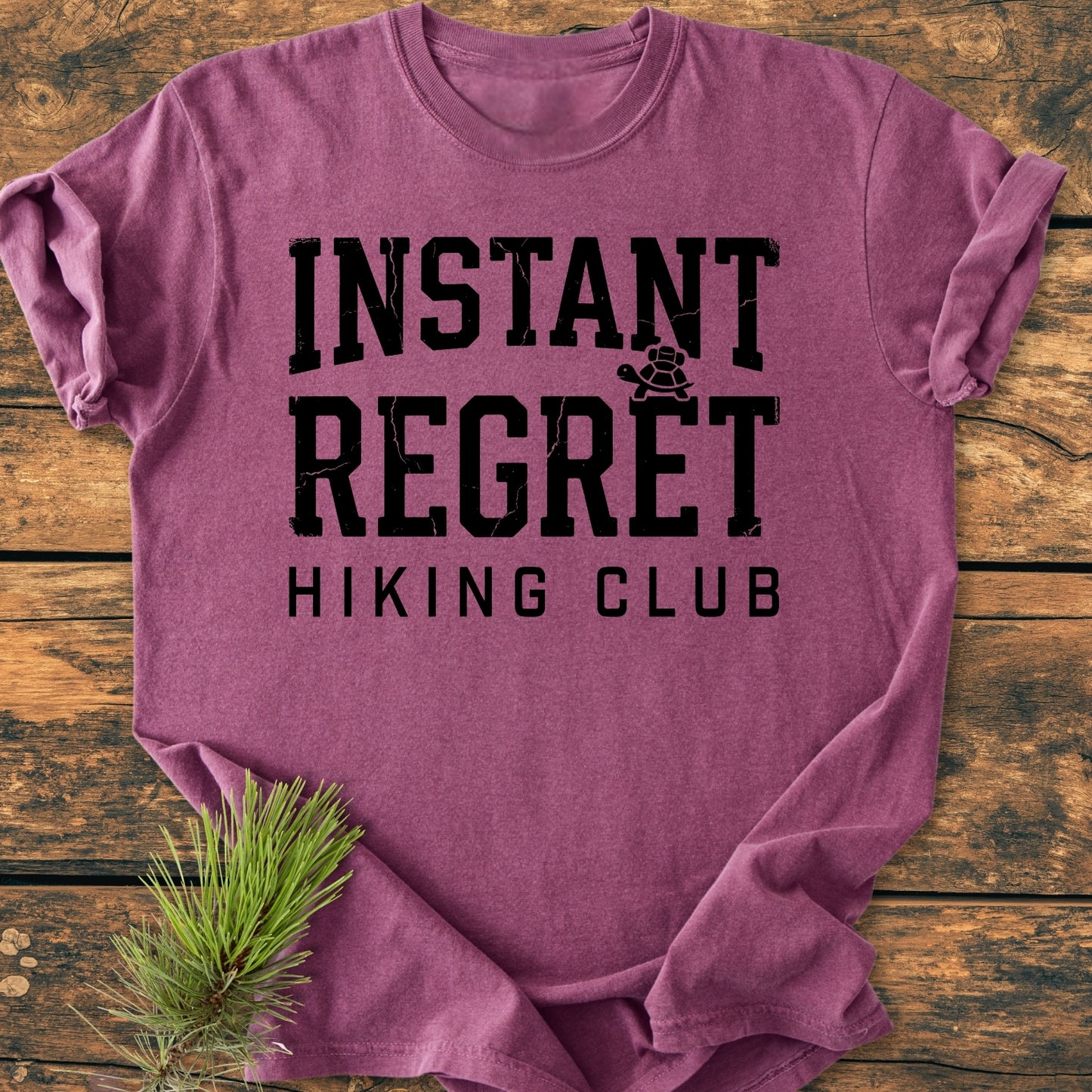 Instant Regret Hiking Club - Vintage Wash Tee: Heather-purple T-shirt with INSTANT REGRET and tortoise icon in distressed black letters, HIKING CLUB below, styled with a pine sprig on wooden surface.