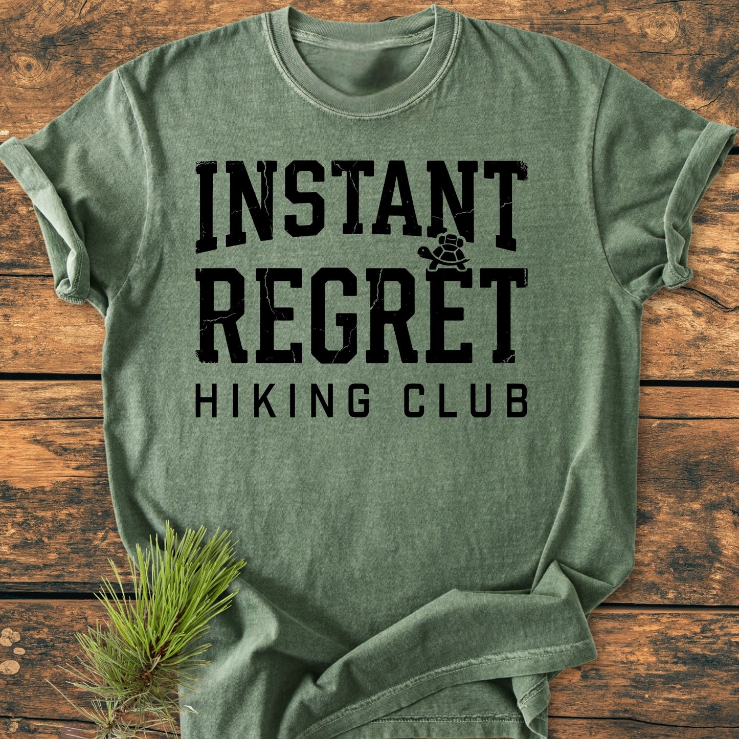 Heather-green Instant Regret Hiking Club - Vintage Wash Tee with black text and tortoise graphic, laid flat on wood with a pine sprig accent, showcasing its relaxed fit and durable stitching.