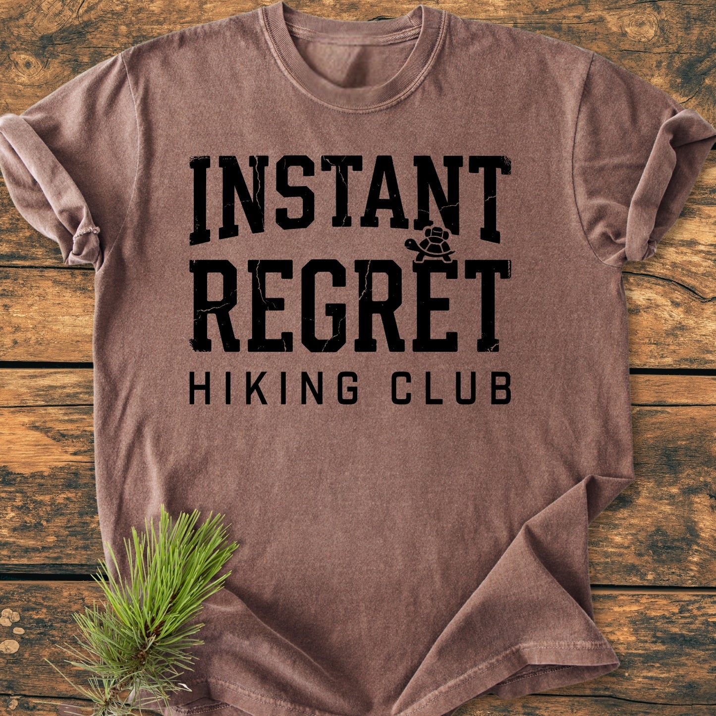 Instant Regret Hiking Club - Vintage Wash Tee folded on wood surface, features INSTANT REGRET with turtle icon and HIKING CLUB below. Pine sprig beside. Soft, durable, relaxed fit, 100% cotton.