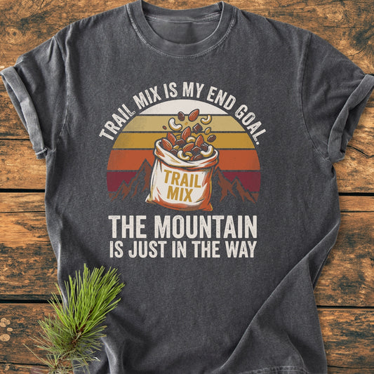 Trail Mix End Goal - Vintage Wash Tee features a retro graphic of spilling trail mix on a dark-gray shirt, laid on wood with a pine branch, highlighting its outdoor theme.