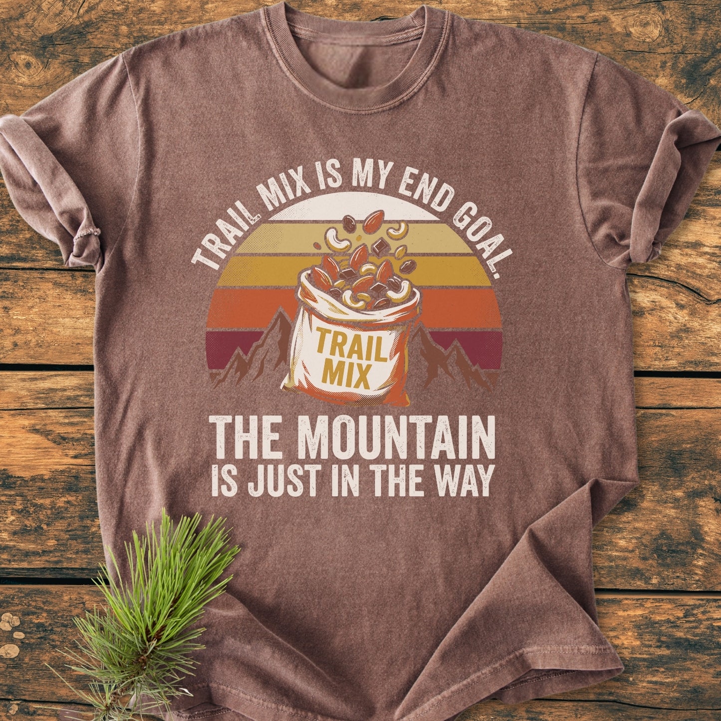 Trail Mix End Goal - Vintage Wash Tee features a retro graphic of trail mix and mountains, showcasing the slogan. Laid flat on wood, highlighting the tee's relaxed fit and durable, soft-washed fabric.