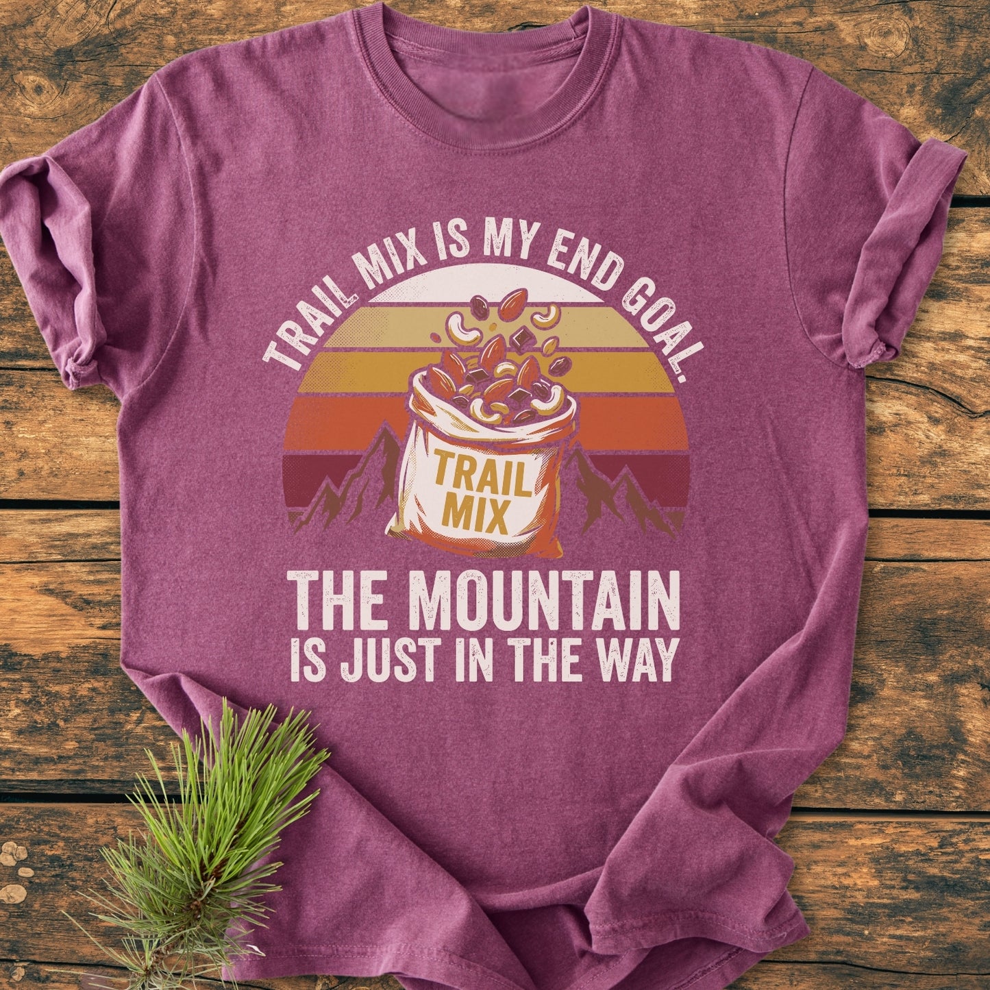 Trail Mix End Goal - Vintage Wash Tee features a retro design with spilled trail mix and mountains, highlighting comfort with 100% ring-spun cotton on a rustic wooden backdrop, accented by a pine sprig.