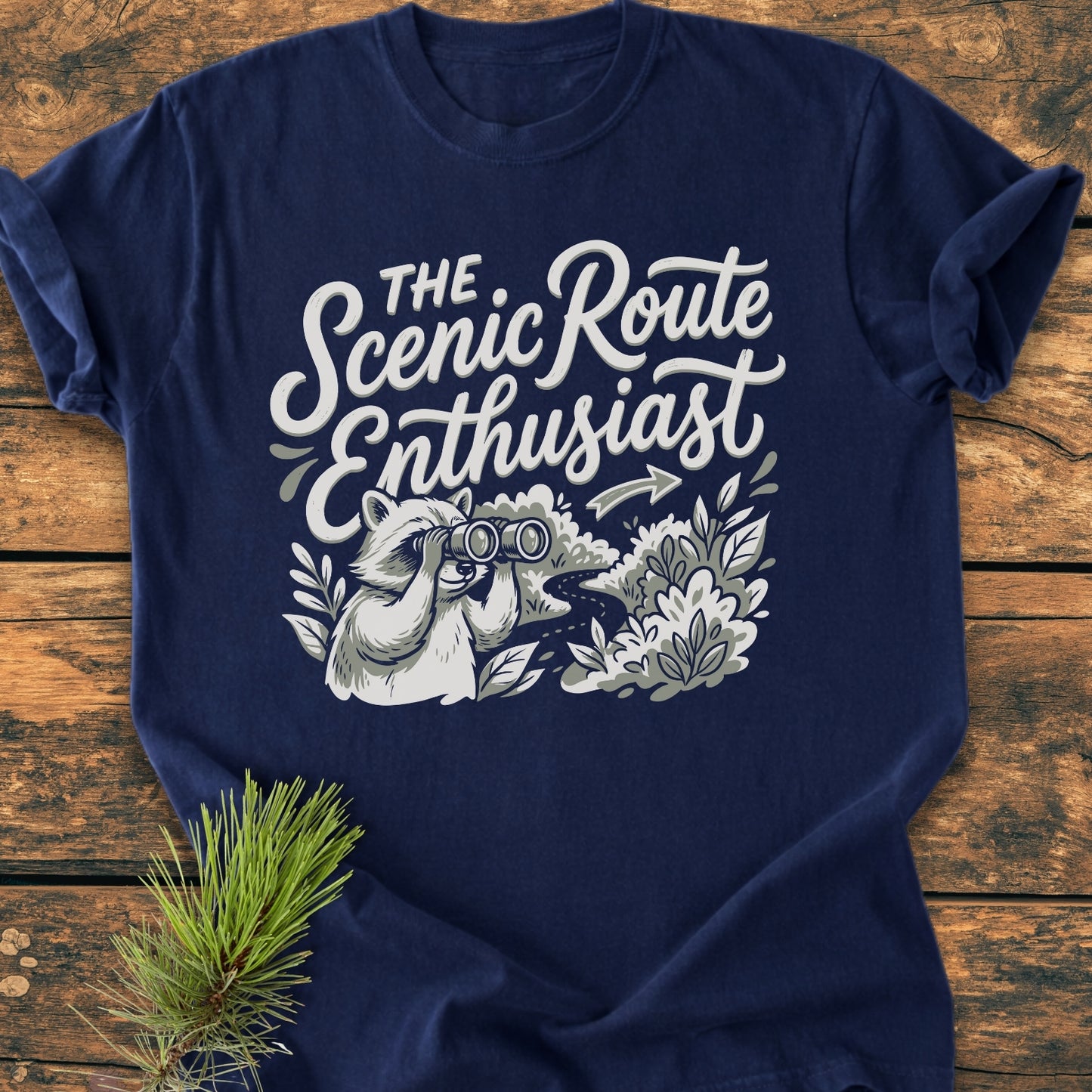 Scenic Route Raccoon - Vintage Wash Tee featuring a playful raccoon graphic with a slogan, laying on a wooden surface with a pine sprig, embodies a cozy, adventure-inspired look.