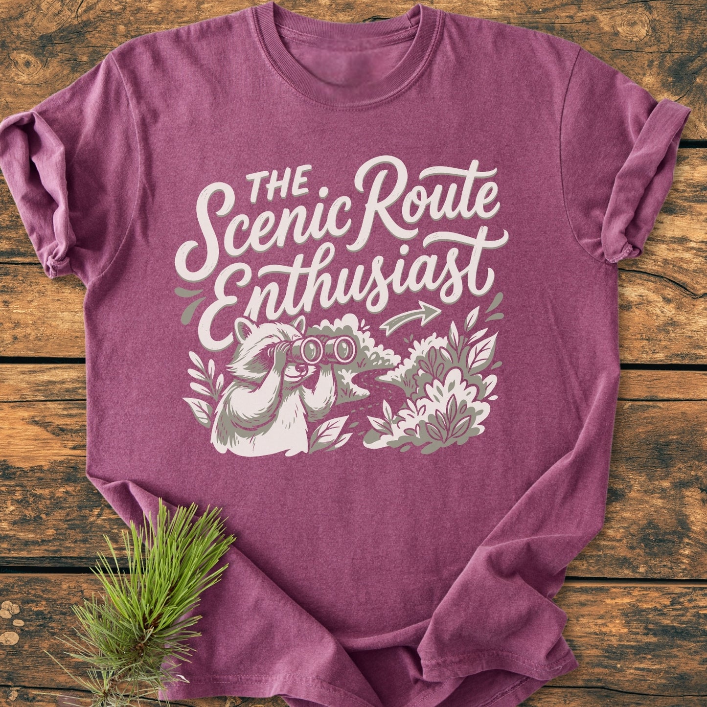 Scenic Route Raccoon - Vintage Wash Tee featuring a cartoon raccoon with binoculars and The Scenic Route Enthusiast slogan. Laid flat on a wooden surface, showcasing its relaxed fit and durable stitching.